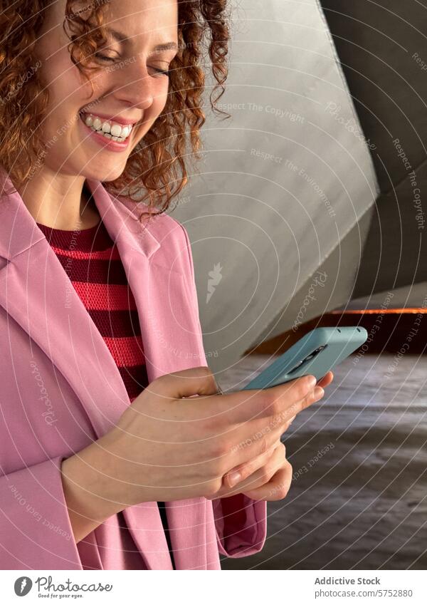 Redhead woman in pink smiling while using smartphone indoors curly hair pink blazer joy communication technology close-up handheld happiness using phone