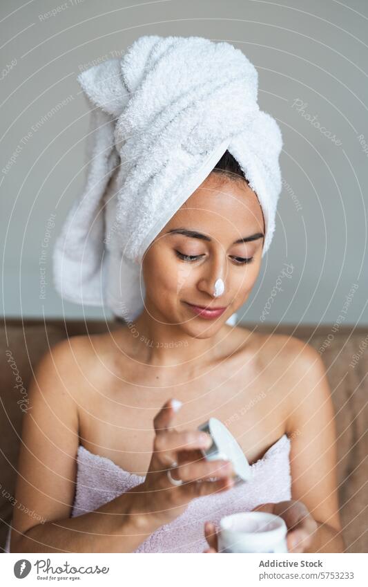 Young woman applying facial cream at home skincare moisturizing self-care beauty routine towel head face young serene wellness health bathroom pampering hygiene