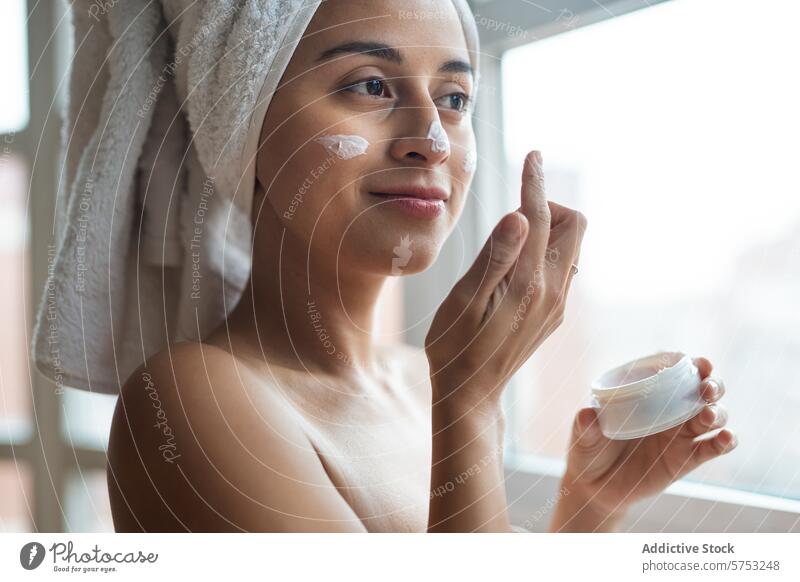 Woman applying facial moisturizer in a cozy room woman skincare cream beauty routine morning towel hair application jar window sunlight self-care bathroom