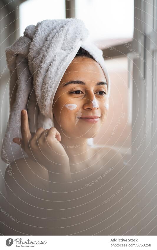 Woman applying facial cream for skincare routine woman moisturizer beauty towel head face serene window morning wellness treatment cosmetic health hygiene