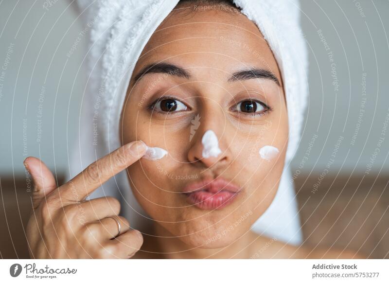 Playful woman applying moisturizer on her face towel headwrap skincare facial cream playful expression beauty routine health lifestyle pampering treatment