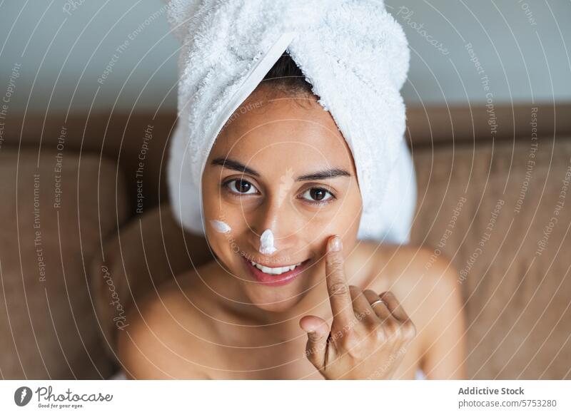 Young woman applying facial cream with a smile skincare moisturizer beauty young radiant towel head face cheerful relaxed routine cosmetic product hygiene