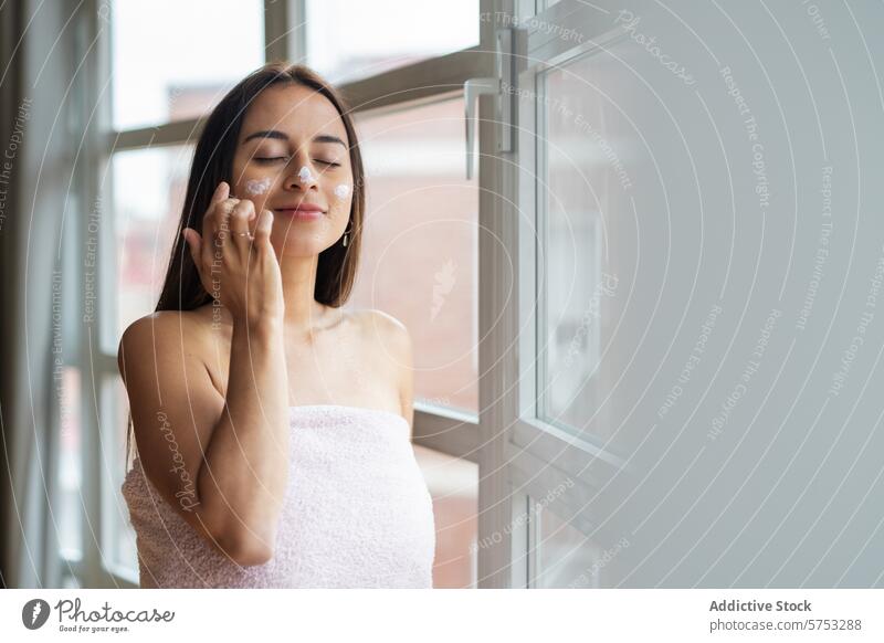 Woman applying facial cream by a window woman skincare moisturizer beauty routine face towel wellness bright hygiene self-care health grooming pampering