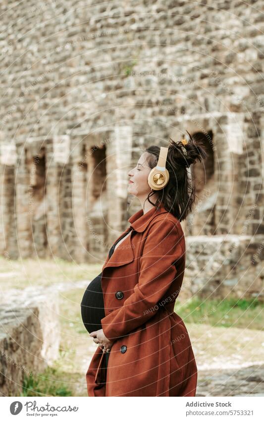 Expectant mother relaxing with music outdoors pregnancy woman expectant headphones relaxation stone wall historical leisure enjoyment peaceful standing coat