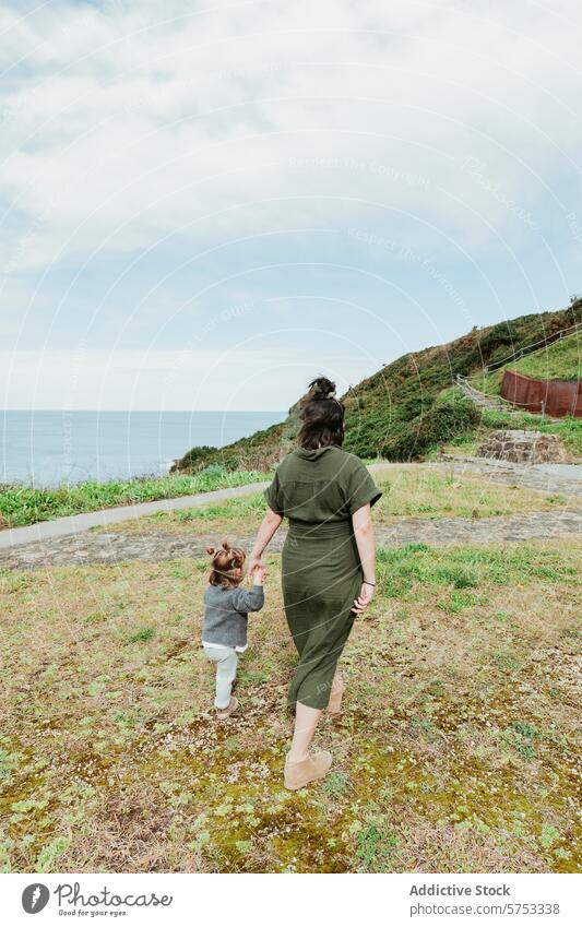 Mother and child walking on coastal path mother hand in hand pathway cloudy sky scenic outdoor family bond parenting toddler nature casual clothing greenery