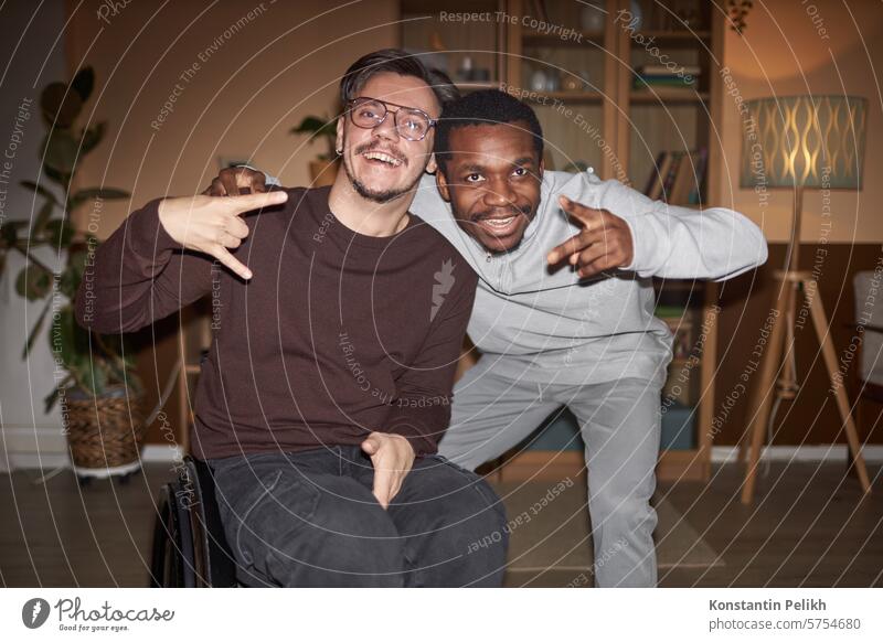 Front view portrait of young man with disability posing with African American friend during house party shot with flash home two wheelchair Black man smile