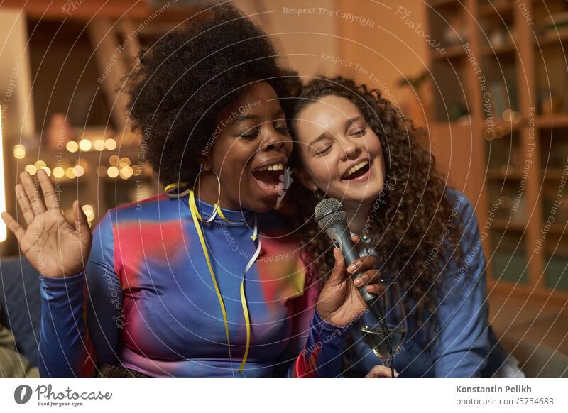 Waist up portrait of two young women singing to microphone and enjoying karaoke during home party people woman Black woman diversity multiethnic song music