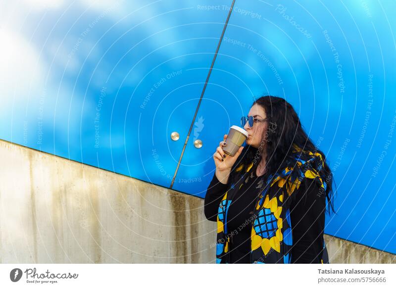Beautiful young brunette woman with long curly hair, dressed in a black, blue and yellow waistcoat, drinks coffee from a paper cup on a background of blue wall
