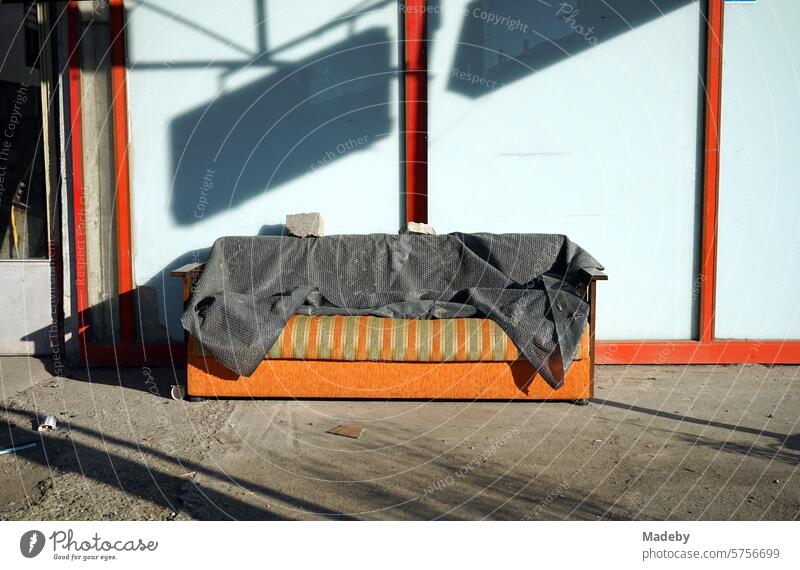 Old discarded striped sofa in green and orange with gray blanket in front of closed store in summer sunshine in Adapazari, Sakarya province, Turkey Veranda