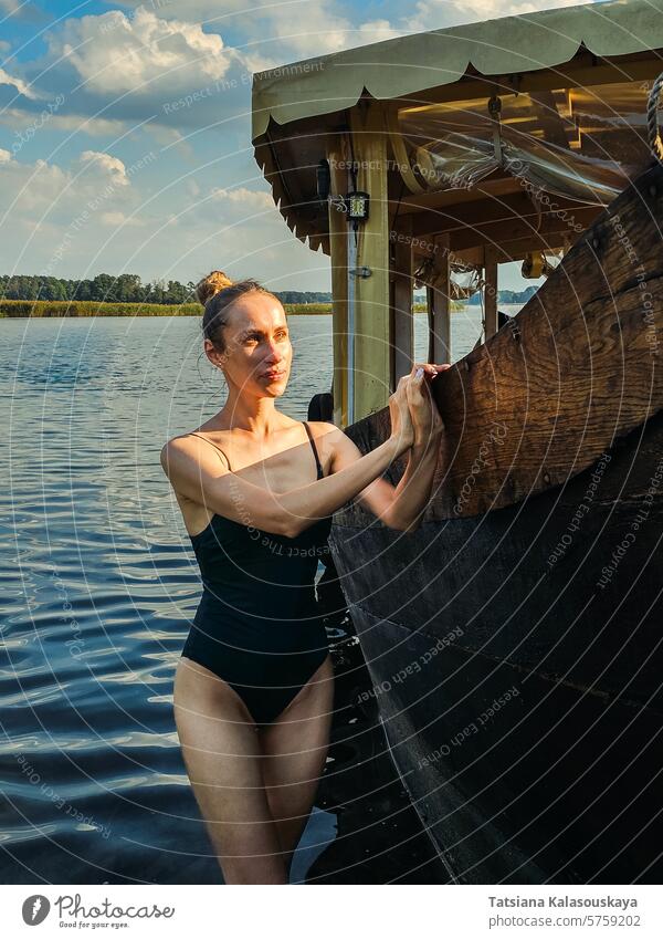 A young slim attractive woman in a one-piece swimsuit is relaxing by the lake and standing leaning against a wooden passenger vintage ship water old college