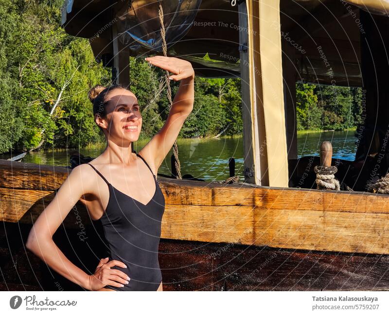 Slender young woman in swimsuit standing near old wooden boat and covering her face from the sun with her palm slender hand sunny slim water vintage smiling