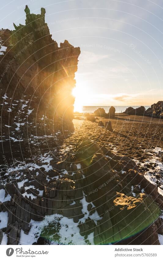 The sun flares around a rugged basalt cliff on an Icelandic beach, highlighting the striking geological formations and the snow's delicate touch golden hour