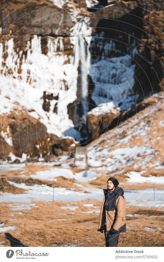 A solitary hiker stands in contemplation, taking in the grandeur of an Icelandic waterfall amidst the contrasting winter landscape man solitude nature travel