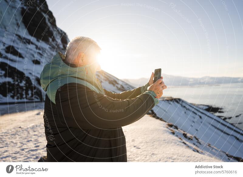 A man captures the moment on his phone, with the winter sun casting a radiant glow over the snowy coastline of Iceland capturing Icelandic shore travel nature