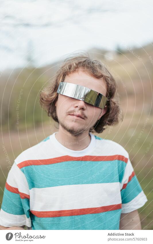 A young man poses in unique, mirrored sunglasses, exuding a cool and contemporary vibe in a natural outdoor setting futuristic fashion casual striped shirt