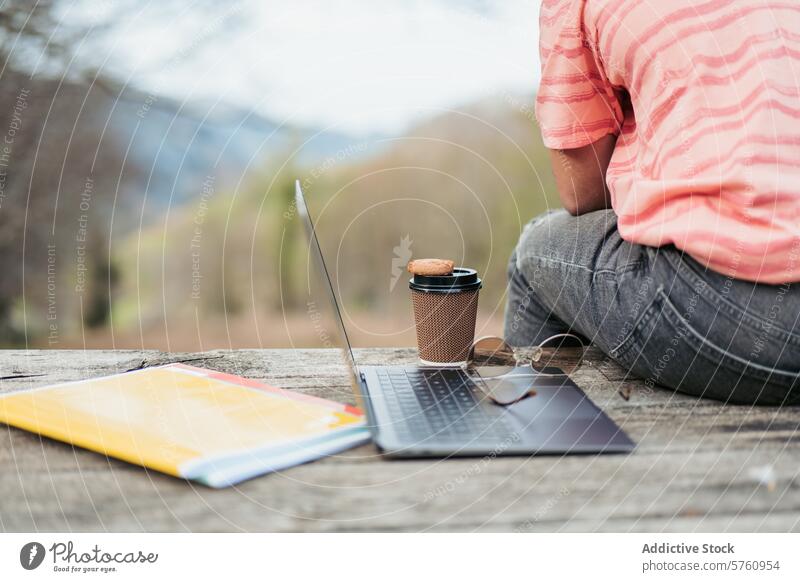 A serene workspace set upon a rustic wooden table outdoors with a laptop, coffee cup, and notepad, surrounded by the tranquility of nature remote study