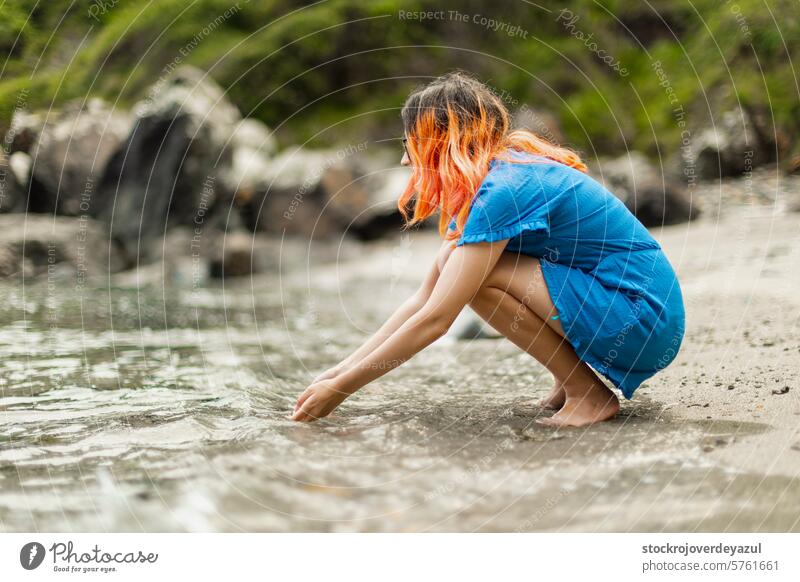 A young girl with orange hair, crouching, touches the water with her hands, while gazing at the seashore beach person summer lifestyle sand nature vacation