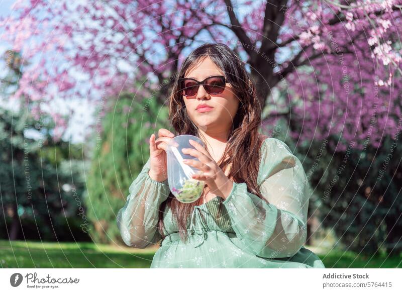 Young woman enjoying a beverage in a blossoming park young sunglasses green dress cup sip pink vibrant outdoor spring nature leisure relaxation tree floral