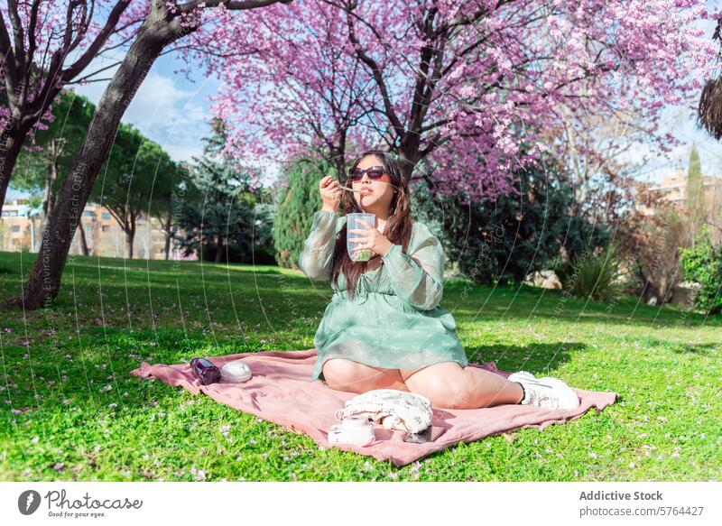 Woman enjoying picnic under cherry blossoms in park woman spring blooming outdoor nature leisure relaxation peaceful pink blanket solo green dress sunglasses