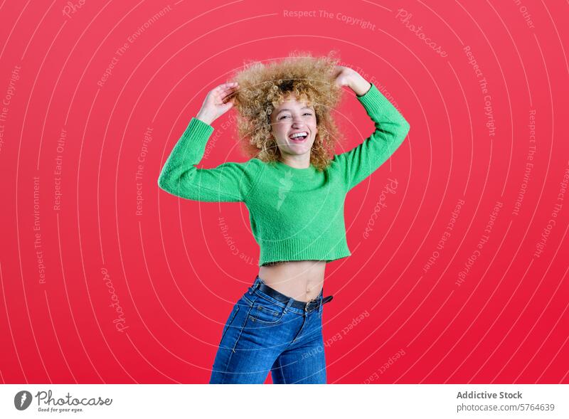 A woman with voluminous curly hair enjoys a playful moment, smiling and touching her hair, clad in a vibrant green sweater and blue jeans, set against a vivid red backdrop