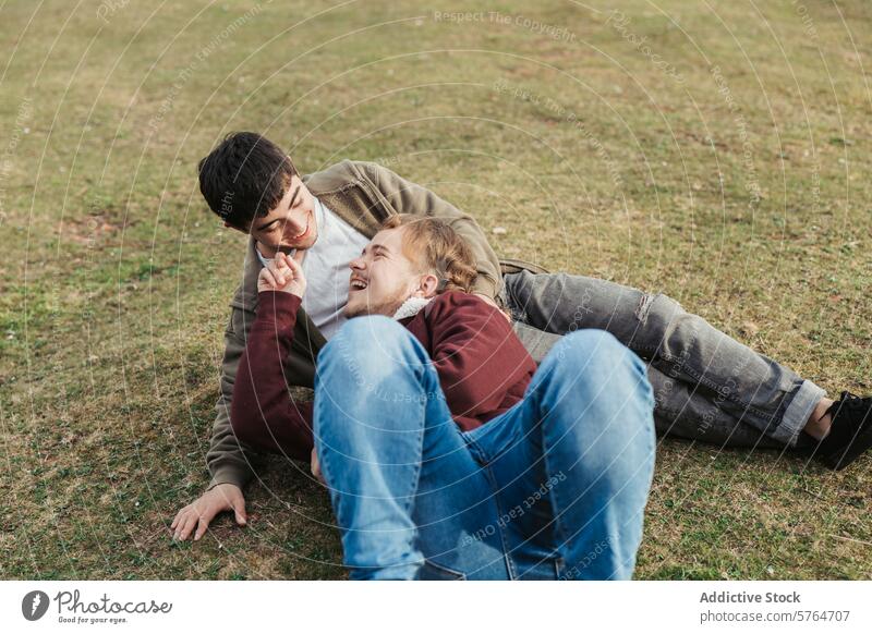 A carefree moment unfolds as one man reclines in the grass, sharing laughter and playful interaction with his partner outdoors field smile casual jean hoodie