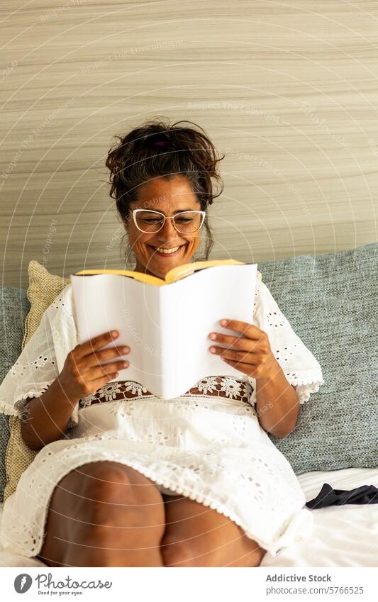 Cheerful middle aged hispanic woman reading on couch book smile cozy white dress enjoyment relaxation leisure hobby literature fiction education glasses resting