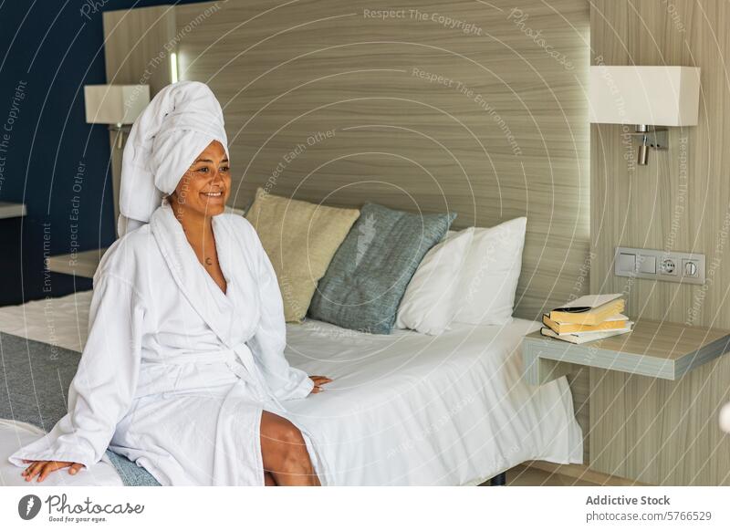 Relaxed middle aged hispanic woman in bathrobe sitting on hotel bed towel leisure comfort serene smile content relaxed bedroom white linens pillow rest