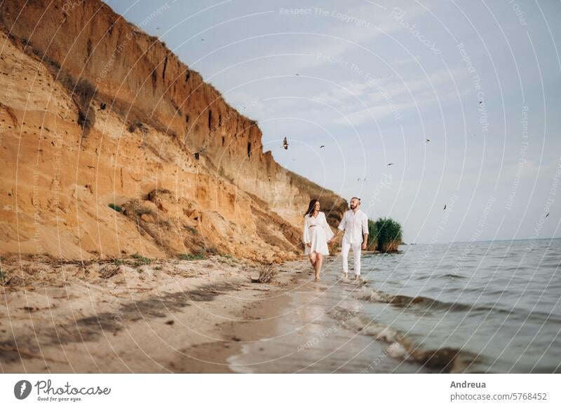 a guy with a girl in white clothes on the seashore husband wife family pregnant coast nearby clay steep cliff mountain water walk sunset light warm summer birds
