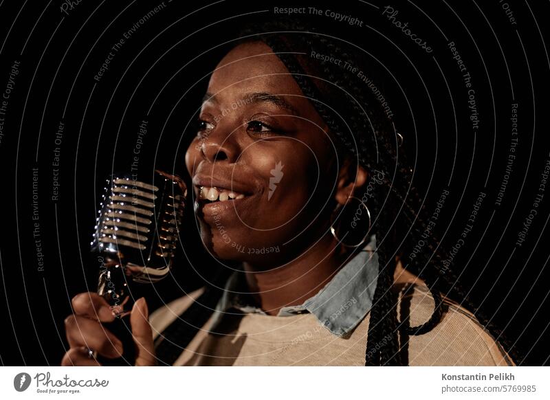 Closeup portrait of smiling African American woman holding microphone while performing on stage with black background copy space sing song concert live music