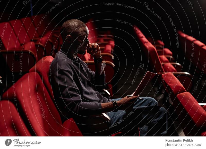 Side view portrait of smiling Black man sitting in audience and reading script in low light at theater copy space artist performer actor rehearse text lines