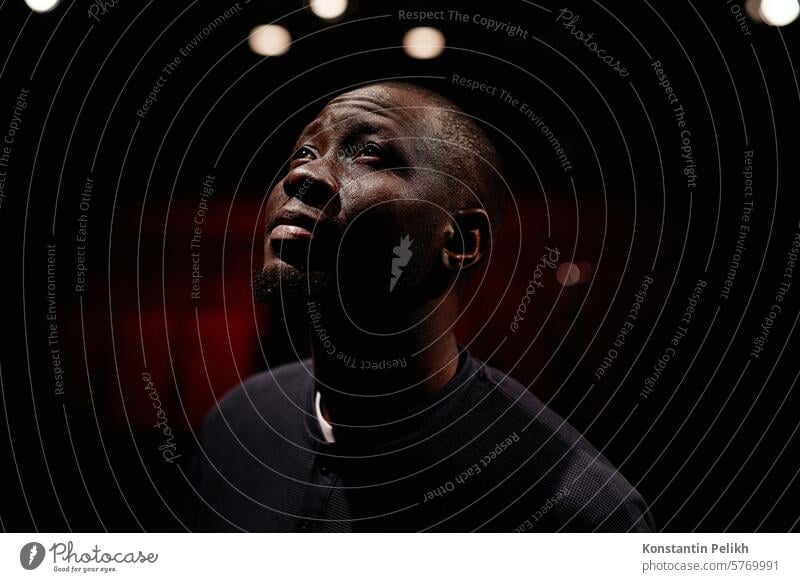 Dramatic portrait of Black adult man looking up into spotlight while standing on stage in theater with low light copy space dream anticipation artist performer
