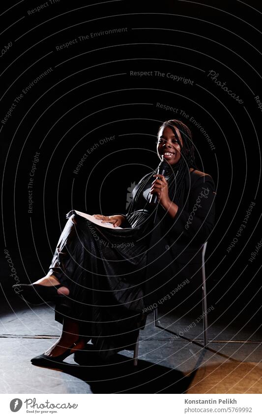 Vertical full length portrait of smiling young woman speaking to microphone on stage while sitting in chair with spotlight stand up comedy talk show comic
