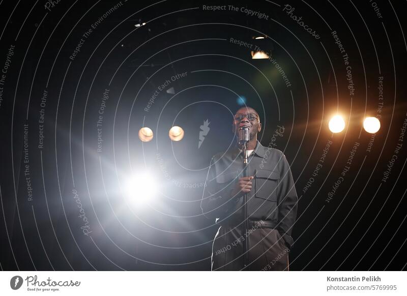 Wide angle shot of African American man speaking to microphone standing on stage with spotlight and smiling copy space stand up comedy talk show comic comedian