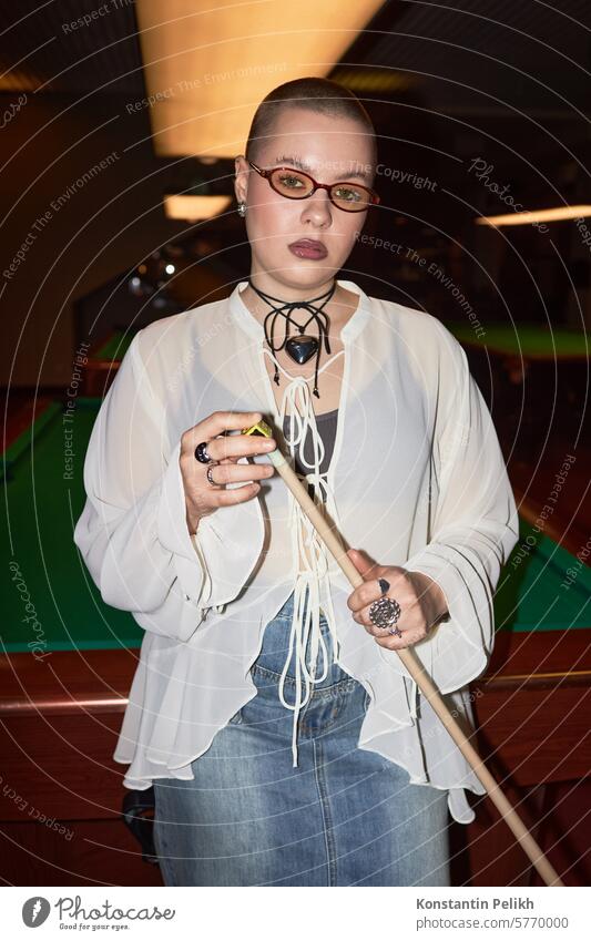 Vertical portrait of bald young woman holding cue stick standing by billiards table in nightclub and looking at camera shot with flash pool girl play game