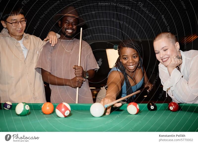 Multiethnic group of smiling young people playing pool together with Black woman hitting ball with cue stick billiard girl game hold aim shot friend table smile
