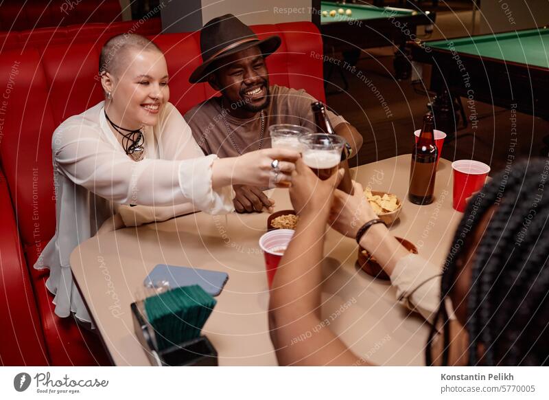 Multiethnic group of friends clinking beer glasses and cheering sitting at diner table cafe people drink cup toast man woman girl smile happy African American