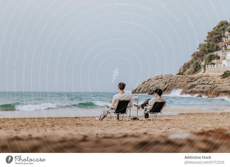 Tranquil beach scene with two people relaxing by the sea chair wave shore sand individual unwind rocky hill backdrop tranquil serene coast leisure summer
