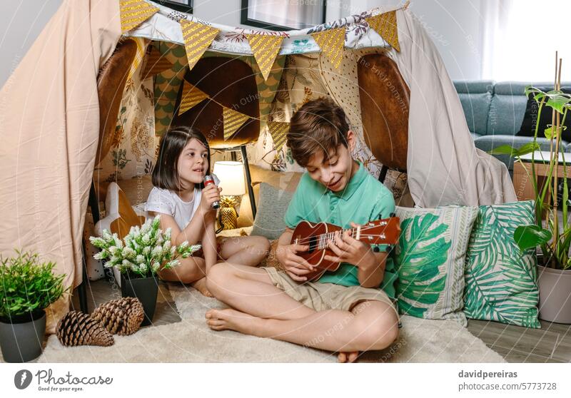 Happy children playing a ukulele and singing on handmade shelter tent in living room at home happy guitar music teepee camping having fun diy indoor homemade