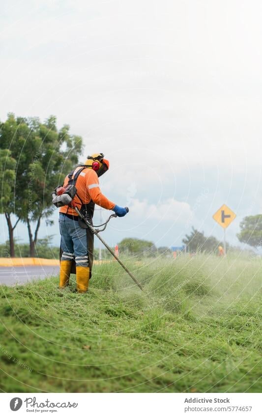 Landscaper trimming grass with a weed scythe landscaper safety gear roadside hearing protection uniform orange tall work worker labor outdoor maintenance
