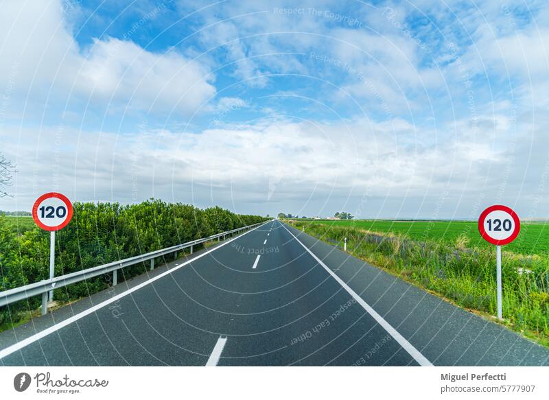 View of a straight highway that disappears into the background bordered by green fields under a blue sky with clouds, with two vertical traffic signs with the limit of 120 km/h. Landscape.