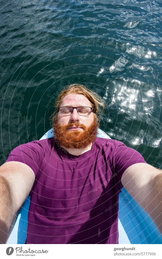 Man with a red beard and glasses lying at the head of a rowing boat and enjoying the sun. Behind him is a lake and the reflection of the sun. The man is wearing a purple shirt, his arms are out in front. Perhaps he is taking a photo of himself.