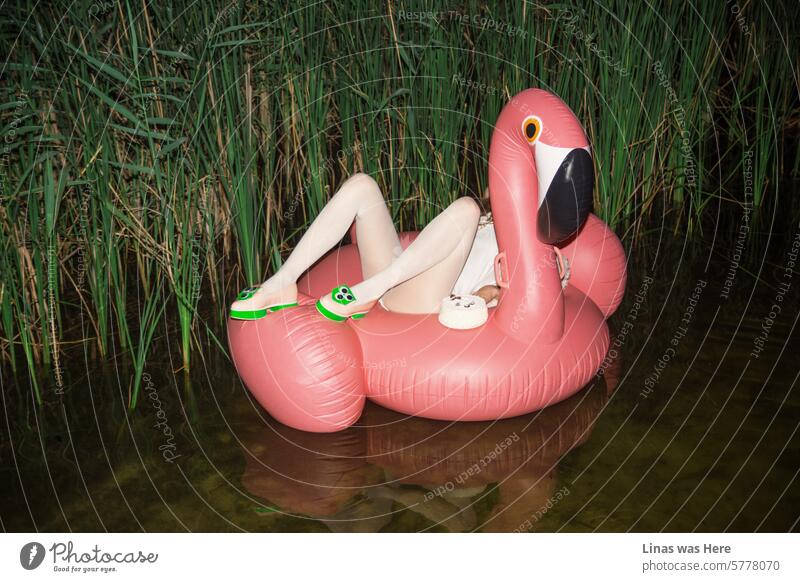 A girl in a white outfit with pretty long legs relaxes on this pink flamingo at night. She has a cake next to her while floating in a lake. Some water reflections, avant-garde shoes, and a chilled atmosphere.
