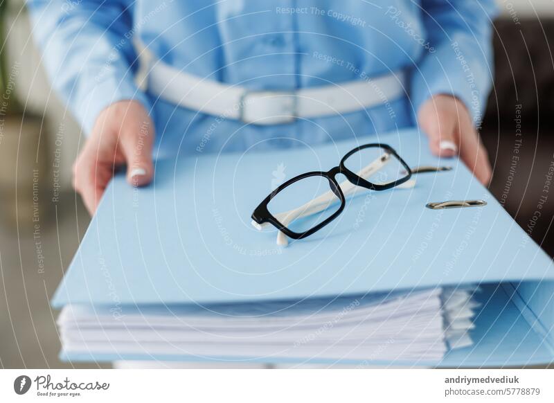 Cropped shot of unrecognisable business woman in blue formal shirt holding full folder of financial documents, selective focus on eyeglass, empty copy space place for advertising text message
