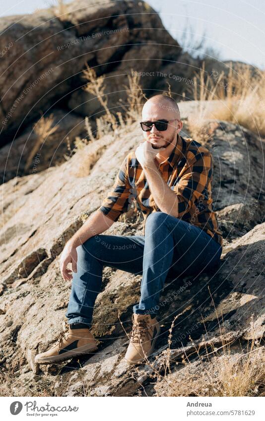 bald guy with a beard in jeans warm shirt and trekking shoes boots glasses see stand wait look into the distance against the background stones granite brown