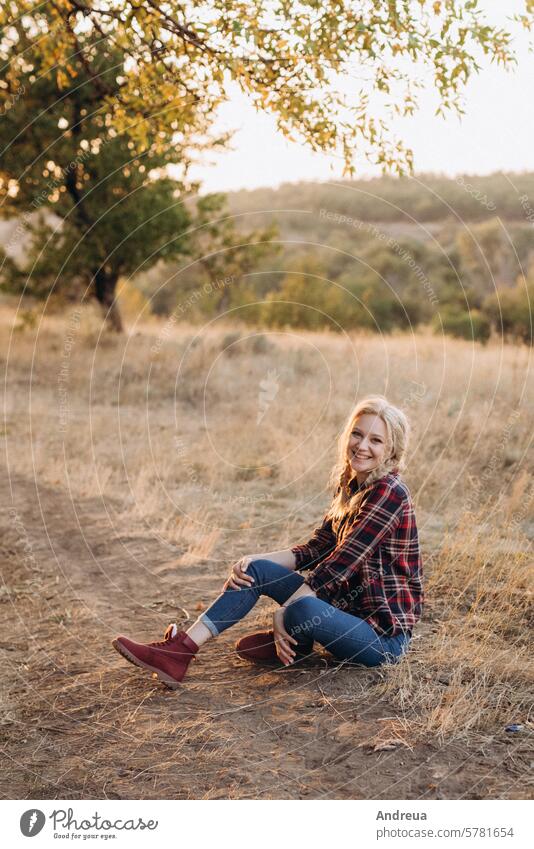 blonde with pigtails in a shirt, jeans, red shoes smiling girl laughs ground grass sunset autumn summer winter boots braids image model joy sit blue brown
