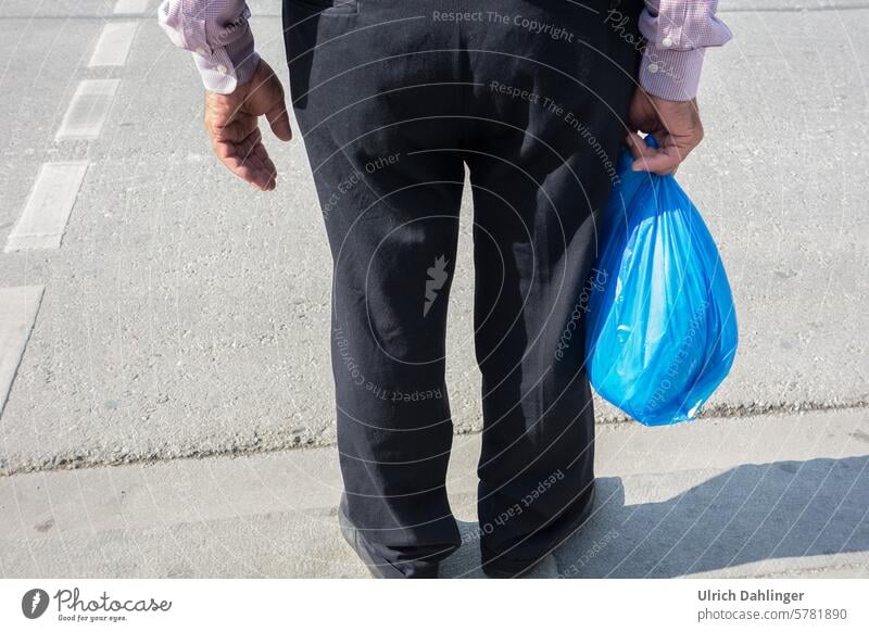 Person standing on the side of the road with a blue plastic bag in his hand, the lower body with black trousers and the hands are shown. Wait Shopping blue bag