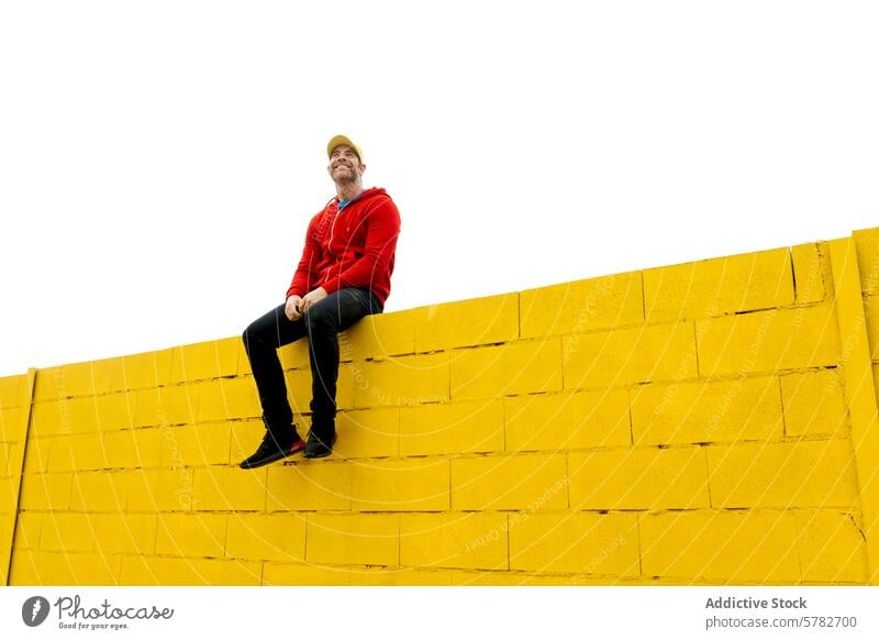 Man in red hoodie sitting on a high yellow wall man casual attire happy joy vibrant color contrast brick white background freedom lifestyle cheerful smiling