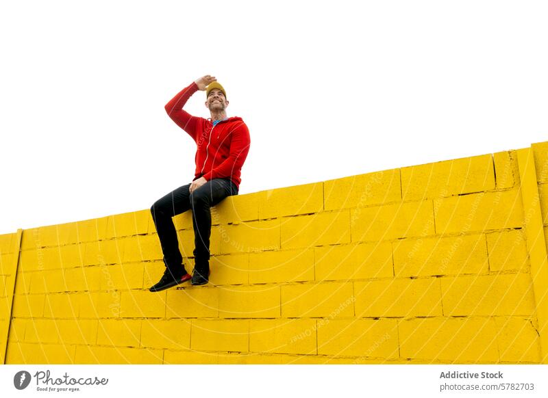 Man sitting on a high yellow wall shielding eyes man sun cheerful red hoodie vibrant bright color outdoor casual looking protection hand gesture sky seated male
