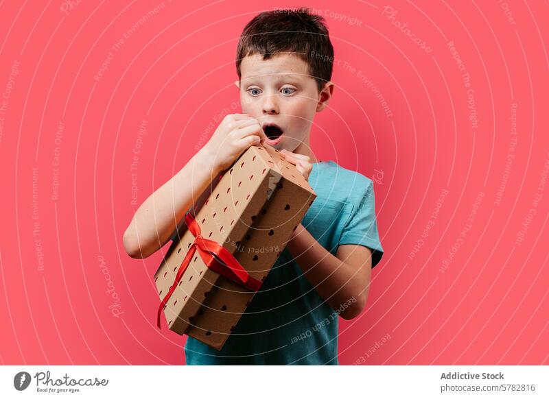 Surprised young boy holding a gift against pink background surprise present red ribbon box child expression astonished excited festive birthday celebration