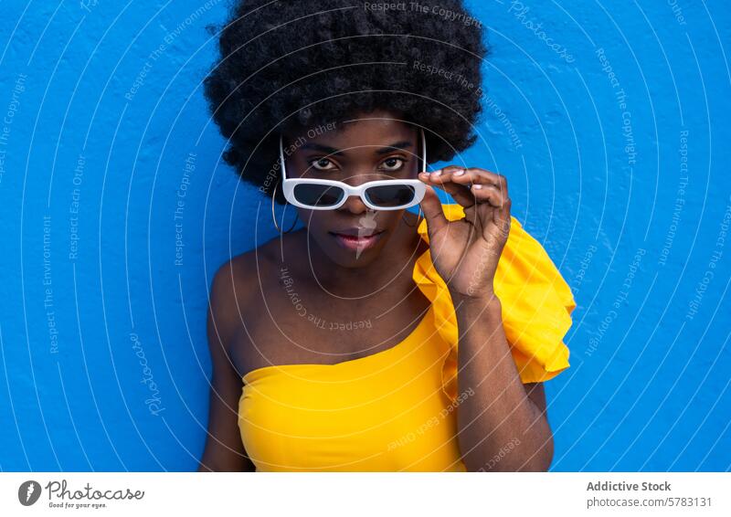 Stylish Afro Woman Posing Against Blue Wall woman afro lifestyle fashion confidence pose yellow dress white sunglasses blue background vibrant chic stylish